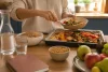 Une personne sert des légumes rôtis avec des céréales, des légumineuses et des fruits sur une table.