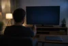 Un homme pointe une télécommande vers un téléviseur à l’écran noir dans un salon.